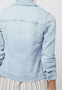 Chaqueta de mezclilla azul claro con mangas remangadas, llevada sobre una falda de rayas blancas y negras, vista desde atrás.