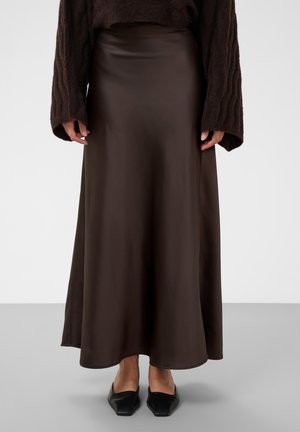 Persona che indossa una gonna lunga in satin marrone scuro, un maglione scuro a maniche lunghe con trama e scarpe nere con punta a punta, in piedi su un pavimento grigio.