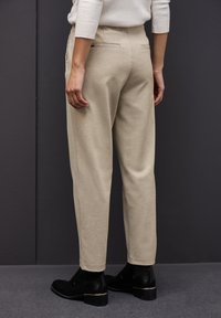Pantalons beige sur mesure en tissu doux, avec une texture lisse, deux poches arrière, associés à des bottines noires avec des accents dorés.