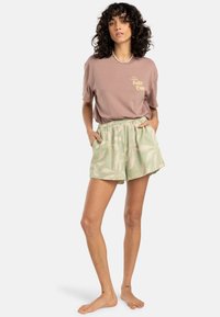 Lichtbruine t-shirt met de tekst "Surf Camp" in het geel, gecombineerd met groene shorts met een palmbladpatroon en een elastische tailleband.