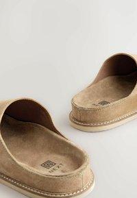 Beige ruskinds slip-on sko med en flad, struktureret sål. Har en rund åben bagdel og syede detaljer langs kanterne. Brandlogo synligt.