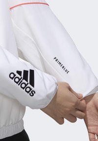Hvid jakke med sort Adidas-logo på ærmet og "PRIMEBLUE" tekst, samt en kontrasterende koralfarvet linje langs skuldersømmen. Letvægtsmateriale.