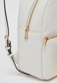 Sac à dos en cuir blanc avec fermetures éclair dorées, finition texturée et bandoulière ajustable. Comprend une poche latérale et une forme arrondie.