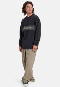 Ciemnoniebieska bluza z pionowym beżowym paskiem z logo, w parze z jasnobeżowymi spodniami cargo i czarnymi sneakersami z białymi podeszwami.