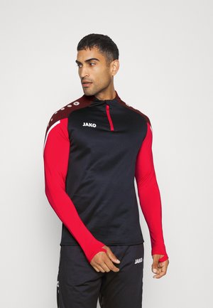 Schwarzes athletisches Langarmshirt mit roten Ärmeln und Schultereinfassungen, ausgestattet mit einem halben Reißverschlusskragen und dem JAKO-Logo auf der Brust.