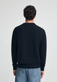 Czarny sweter z długim rękawem z ribbowanym dekoltem i mankietami. Noszony z jasnoniebieskimi dżinsami, prezentującymi gładką, jednolitą teksturę materiału.