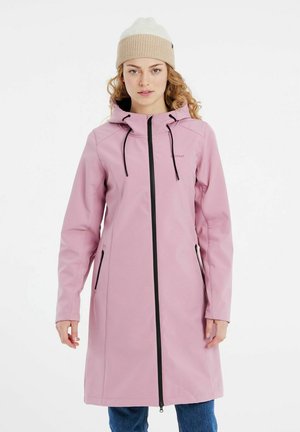 Manteau imperméable rose avec une capuche, des manches longues, une fermeture éclair noire et des poches avant. Le modèle porte un bonnet en tricot beige.