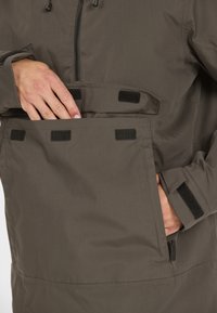 Oliivinvihreä takki, jossa on vetoketjukiinnitys, iso etutasku tarrakiinnityksellä ja säädettävät hihansuut. Materiaali vaikuttaa teksturoidulta ja kestävältä.