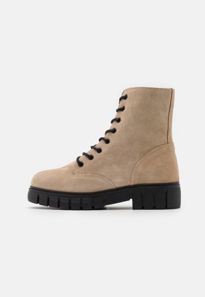 Bottine beige en daim à lacets avec semelle en caoutchouc noir épaisse et bout rond, vue de profil sur fond blanc.