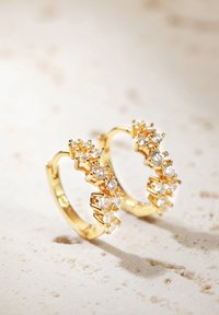 Pendientes de aro dorados adornados con pequeñas piedras transparentes, que presentan un diseño texturizado y una silueta curva, reposando sobre una superficie de piedra clara.