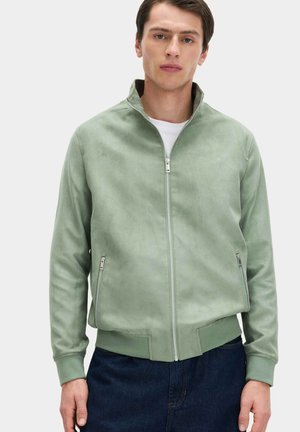 Junger Mann trägt eine hellgrüne Wildlederjacke mit Reißverschluss und seitlichen Reißverschlusstaschen über einem weißen Hemd und dunkelblauen Jeans.