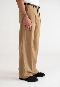 Beige wijde broek met een gladde textuur, voorzien van een voornaad en een bijpassende leren riem in bruin, gecombineerd met zwarte chunky schoenen.
