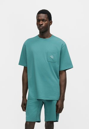 Homme portant une chemise oversize manches courtes bleu sarcelle avec poche poitrine et un short assorti, debout devant un fond clair uni.
