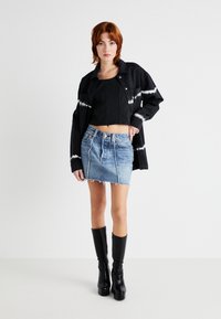 Jachetă scurtă neagră cu model alb de tie-dye, bluză neagră, fustă denim mini cu tiv desfăcut și bocanci negri până la genunchi.