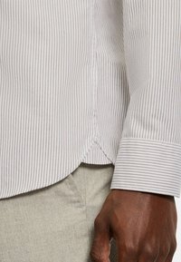 Hellgrau gestreiftes Hemd mit dezenten braunen Linien, abgerundetem Saum und geknöpften Manschetten. Nahaufnahme der Stoffstruktur und einer Hand, die das Hemd greift.