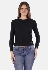 Maglione nero a maniche lunghe con lavorazione a costine, scollo rotondo, design aderente e dettaglio del logo all'orlo. Indossato con jeans di denim blu.