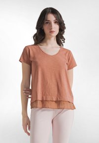 Roestkleurig T-shirt met korte mouwen, V-hals en laagjesontwerp met een transparante zoom. Draagt lichte roze nauwsluitende broek.