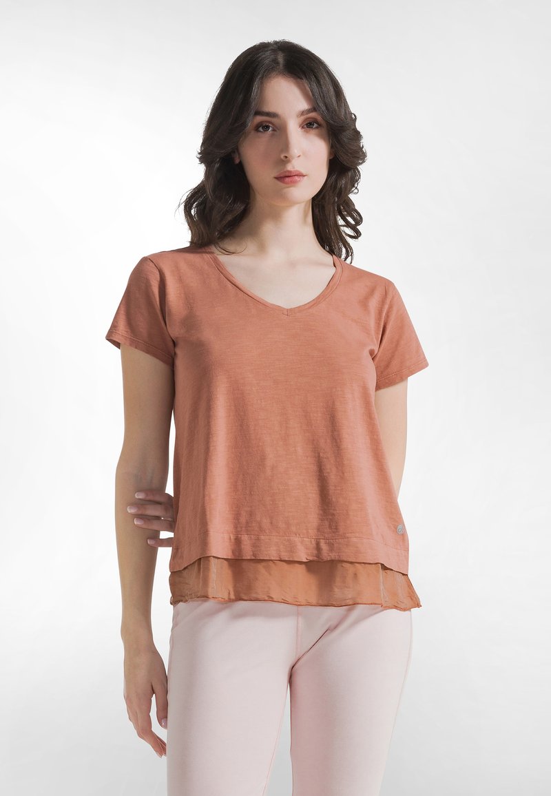 Roestkleurig T-shirt met korte mouwen, V-hals en laagjesontwerp met een transparante zoom. Draagt lichte roze nauwsluitende broek.