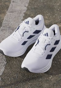 Paire de chaussures de sport blanches avec des bandes latérales bleu marine sur une surface en asphalt avec une ligne peinte.