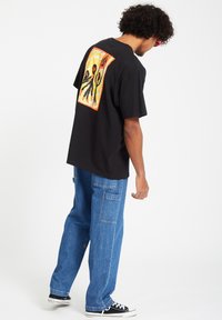 Black cotton t-shirt featuring a colorful graphic on the back with abstract figures. Loose fit, paired with light blue denim pants and black shoes.