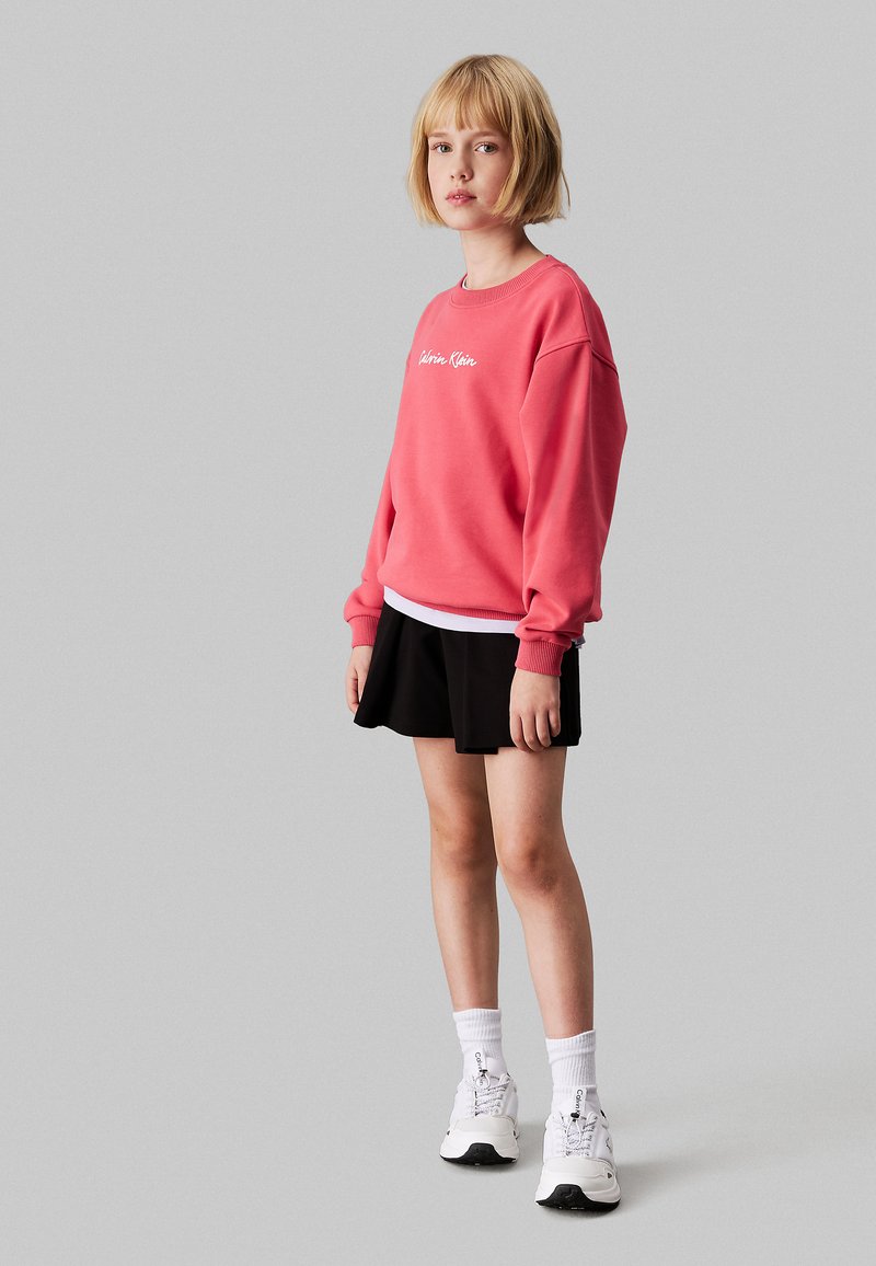 Felpa rosa con logo bianco, abbinata a una gonna plissettata nera. Calze bianche e sneakers completano l'outfit. Capelli corti.