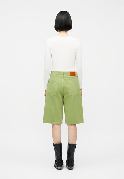 Lühikeste mustade juustega inimene, kes kannab valget pika varrukaga salli, rohelist põlvikutega pükse, millel on tagataskud, ja musti vasikasäärseid saapaid, seistes seljaga vaataja poole.