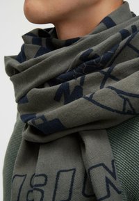 Gray scarf wrapped around the neck, featuring a blue geometric pattern and soft texture; displayed against a green knitted background.
