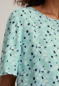 Lichtblauw t-shirt met korte mouwen, voorzien van een abstract patroon in zwart en groen, met verschillende vormen zoals bladeren en geometrische figuren.