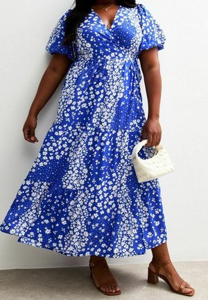 Mujer con un vestido cruzado azul con flores y mangas abullonadas, sosteniendo un pequeño bolso blanco tejido y llevando sandalias marrones con tacón grueso.