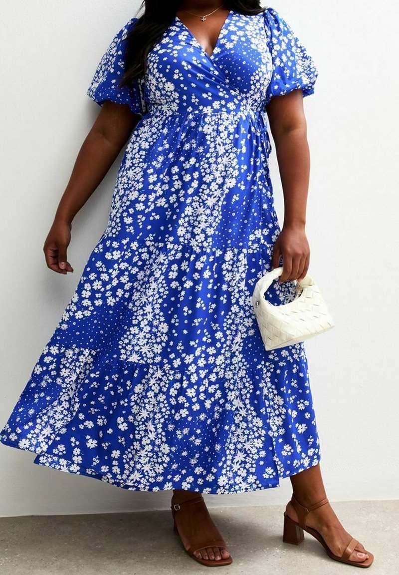 Femme portant une robe portefeuille bleue à motifs floraux avec des manches bouffantes, tenant un petit sac à main blanc tissé et portant des sandales marron à talons blocs.