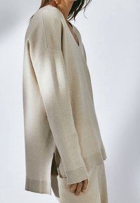 Pull oversize beige avec un col en V profond, des poignets côtelés et des fentes latérales. Texture douce et coupe décontractée.