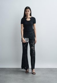 Mujer con una camiseta negra de manga corta con un recorte delantero, pantalones negros de encaje, tacones negros y sosteniendo un pequeño bolso de mano metálico.