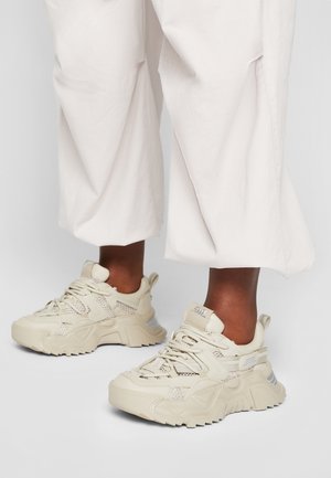 Beige chunky sneakers met mesh- en lederen panelen, gedragen met losse, witte cropped broeken op een donkere huid.