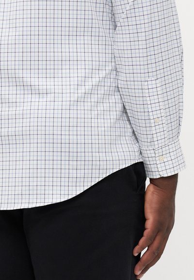 Chemise blanche à motif quadrillé bleu, à manches longues, avec des poignets boutonnés, ourlet arrondi, et en tissu lisse et léger. Portée avec un pantalon sombre.