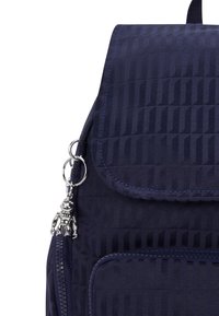 Mörkblå ryggsäck med en texturerad design, med klaffstängning, zippered fickor och en silverfärgad astronautcharm fäst vid en nyckelring.