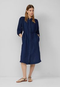 Vestito blu navy, di lunghezza al ginocchio, in tessuto leggero con chiusura a bottoni, maniche a tre quarti, vita legata e tasche laterali, abbinato a sandali color beige.