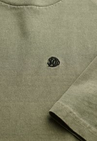 Zielona oliwkowa bawełniana koszulka z okrągłym dekoltem. Posiada czarne haftowane logo. Miękka w dotyku, z krótkimi rękawami i luźnym krojem.