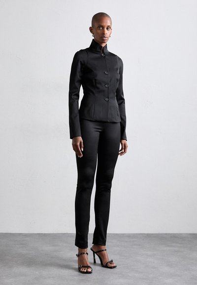 Black fitted jacket with high collar and button closure, paired with sleek black trousers and strappy high-heeled sandals. Smooth fabric texture.