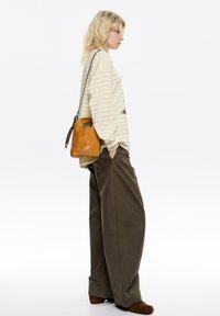 Skórzana torba na ramię w odcieniu brązowym, z zaciągnięciem, noszona z kremową, w paski, długą bluzką z długim rękawem oraz oversize'owymi brązowymi spodniami.