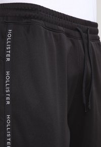 Svarta sportshorts i slät tyg, med dragsko i midjan, sidofickor och vit "HOLLISTER"-logotyp längs sidan.