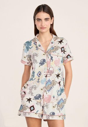 Mujer joven con camisa de manga corta abotonada y pantalones cortos a juego con patrones abstractos coloridos de animales y estrellas, manos en los bolsillos.