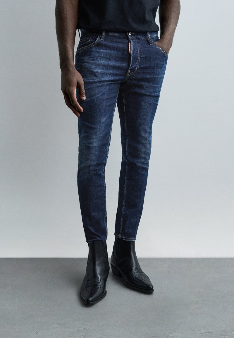 Homme portant un jean slim bleu foncé et des bottes noires à bouts pointus, debout avec une main dans la poche devant un fond uni.