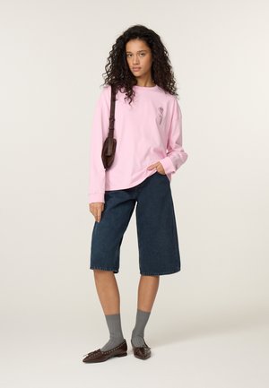 Frau mit lockigem Haar, die ein pinkes Langarmshirt, dunkelblaue knielange Shorts, graue Socken, braune Ballerinas und eine braune Umhängetasche trägt.