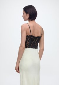 Γυναίκα με κοντά σκούρα μαλλιά που φορά χρυσά σκουλαρίκια, μαύρο κορσέ από δαντέλα και λευκή σατέν φούστα, εμφανίζεται από πίσω πάνω σε λευκό φόντο.