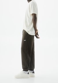 Ljusbeige t-shirt med kort ärm, bruna joggers med elastiska mudd, vita sneakers med marinblå detaljer, modell stående i profil.