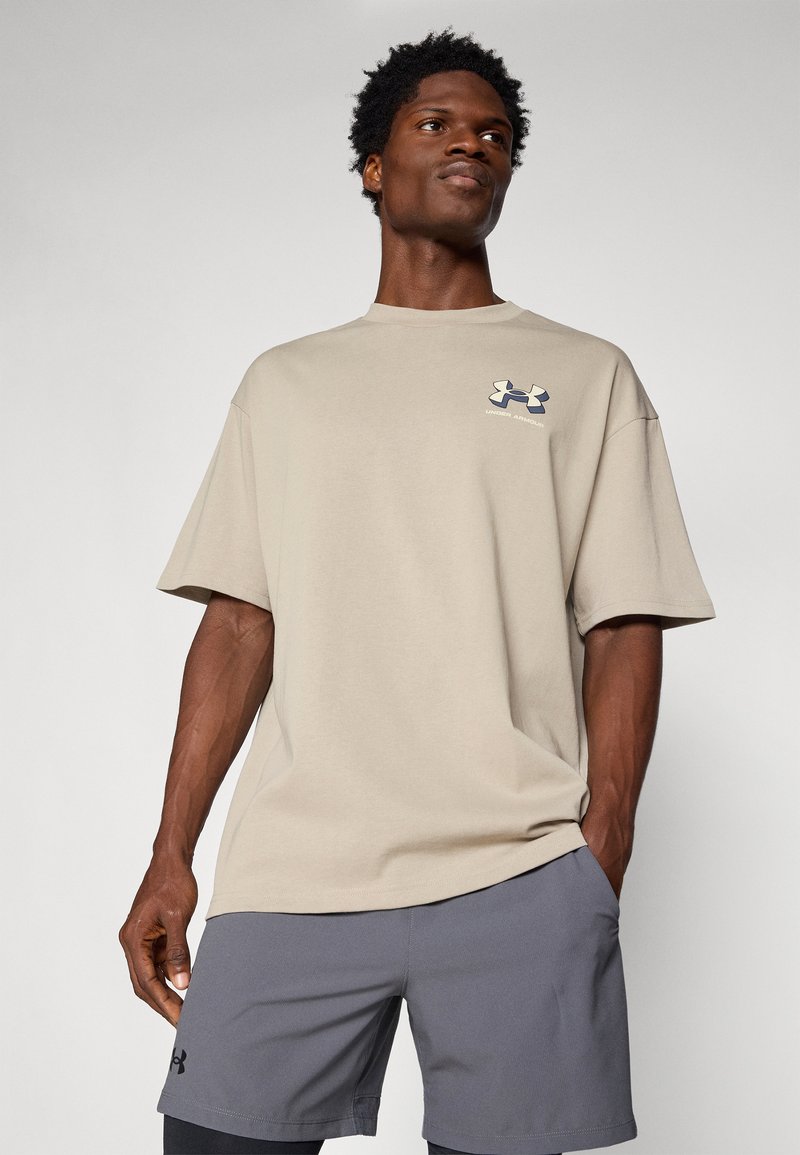 T-shirt beige a maniche corte con una vestibilità rilassata, caratterizzato da un logo Under Armour blu navy sul petto. Indossato con pantaloni corti grigi.