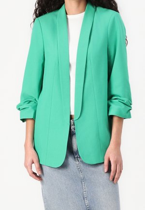 Kobieta w jaskrawozielonym żakiecie z podwiniętymi rękawami, białej prążkowanej bluzce i jasnoniebieskiej spódnicy dżinsowej na jednolitym białym tle.