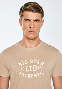 Beežist puuvillast t-särk ümar kaelusega, millel on valge kulunud trükk tekstiga "BIG STAR LTD 1979 AUTHENTIC" rinnal.