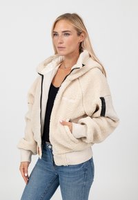 Kremowa kurtka sherpa z kapturem, z przodu na zamek, z prążkowanymi mankietami. Posiada boczne kieszenie i teksturowane wykończenie. Czarne akcenty na rękawach.