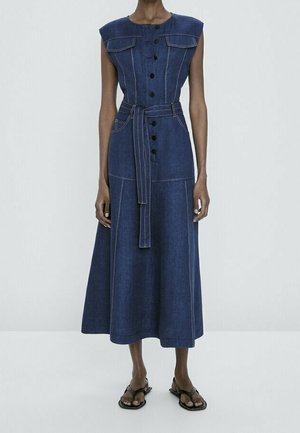 Robe midi en jean bleu foncé sans manches avec boutons sur le devant, ceinture, larges épaulettes et poches, portée avec des sandales noires.